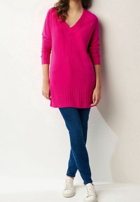Fuchsia knit sweater with a deep V-neck, ribbed hem, and long sleeves, paired with blue jeans and white sneakers.