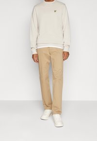 Hombre vestido con pantalones beige, zapatillas blancas y un suéter ligero beige con un pequeño logotipo amarillo en el pecho, de pie frente a un fondo liso.