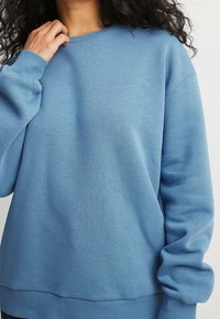 Mujer vistiendo un suéter largo de manga larga y holgado de color azul claro, con una mano cerca del cuello y cabello rizado oscuro visible.