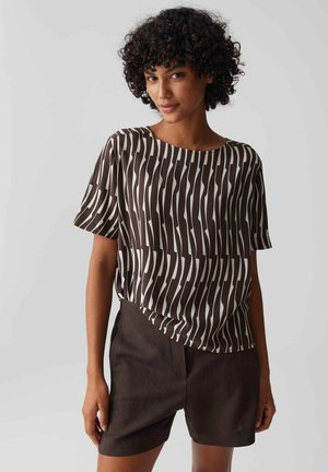 Frau mit lockigem Haar, die ein braun-weiß vertikal gestreiftes Kurzarmtop und dunkelbraune Shorts trägt, steht vor einem einfarbigen Hintergrund.