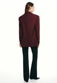 Kvinna i en vinröd blazer och svarta rutiga byxor som står, vänd bort med huvudet vänt åt höger mot en vit bakgrund.