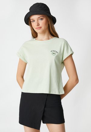 Ljusgrön bomulls t-shirt med uppvikta ärmar, med en smiley-ansikte grafik och texten "Like Feelin’ Good" nära bröstet, ihop med svarta shorts.