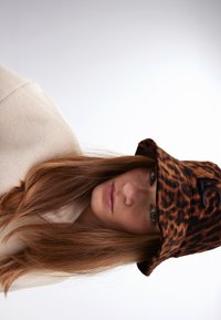 Klobouk s leopardím vzorem, vyrobený z měkkého materiálu, s černým logem jako akcentem. Modelka má na sobě krémový fleece top, s dlouhými vlasy viditelnými.