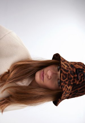 Kuviohattu leopardinprintillä, jossa on pehmeä rakenne ja musta logo. Mallilla on päällä kermaisen värinen fleecepaita ja pitkät hiukset näkyvissä.