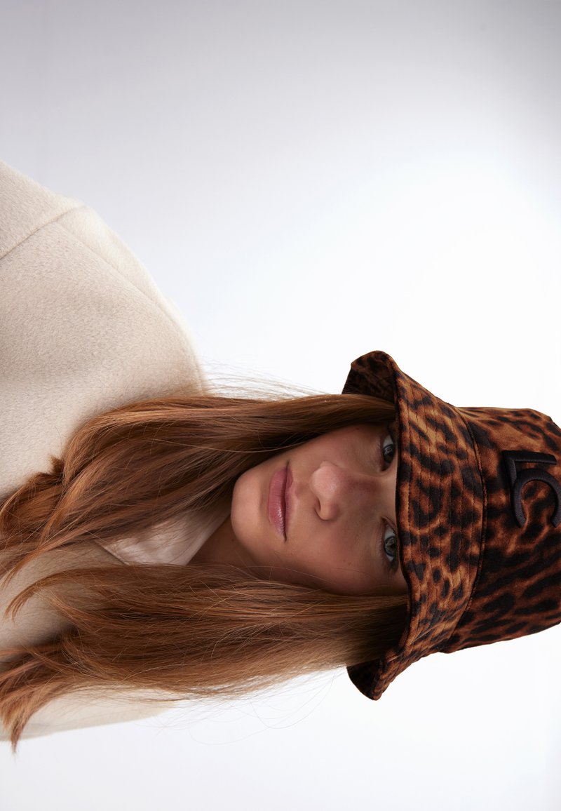 Klobouk s leopardím vzorem, vyrobený z měkkého materiálu, s černým logem jako akcentem. Modelka má na sobě krémový fleece top, s dlouhými vlasy viditelnými.