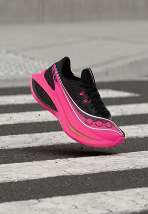 Zwarte en felroze hardloopschoen met dikke zool, in de lucht boven een zebrapad op een asfaltweg.