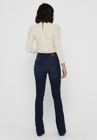 Cream puff-sleeve top with open back, paired with dark blue flared jeans featuring back pockets and a brown leather patch on the waistband.