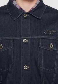Veste en jean foncé avec des boutons en laiton, deux poches poitrine, des coutures beige et une chemise à col à carreaux en dessous.