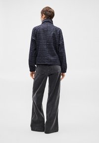 Σκούρο μπλε καρό φλις μπουφάν με ψηλό γιακά, φορεμένο πάνω από γκρίζες φαρδιές παντελόνες. Διαθέτει ελαστικές μανσέτες και απαλή υφή.