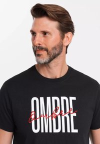 Camiseta de algodón negra con texto en bold blanco "OMBRE" con un acento en cursiva roja. Cuello de tripulación y mangas cortas, textura acanalada.