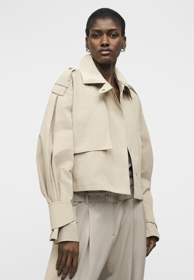 Γυναίκα φορώντας μια μπεζ oversized ζακέτα με φαρδιές μανσέτες και πλισέ μανίκια, συνδυασμένη με ταιριαστά ψηλόμεσα παντελόνια και ζώνη.