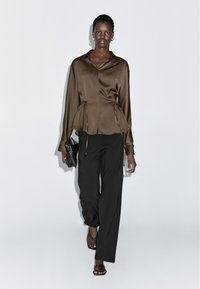 Bruine satijnen blouse met een trekkoord in de taille, gecombineerd met losse zwarte broek. Model houdt een zwarte clutch vast en draagt platte sandalen.