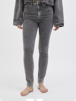 Person iført højttaljede, grå skinny jeans og en matchende cropped denimjakke, stående barfodet på en ensfarvet hvid baggrund.