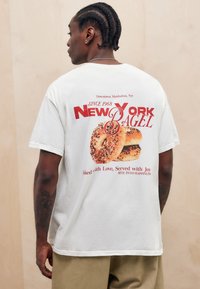 Bāts balts krekls no kokvilnas ar sarkanu grafisku dizainu, kurā attēlotas divas bageles ar tekstu: "New York Bagel" un "Since 1968" aizmugurē.