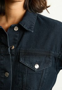 Chaqueta de mezclilla oscura con botones de presión plateados, un cuello puntiagudo y un bolsillo en el pecho con sutiles detalles de costura.