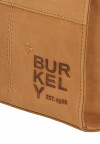 Tan leren tas met gestikte details en een gegraveerd "BURKELY EST. 1988" label voor branding. Zachte textuur met afgeronde hoeken.