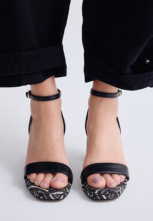 Pies que llevan sandalias negras con tiras en el tobillo, suelas estampadas y pantalones negros de pierna ancha recortados por encima del tobillo.