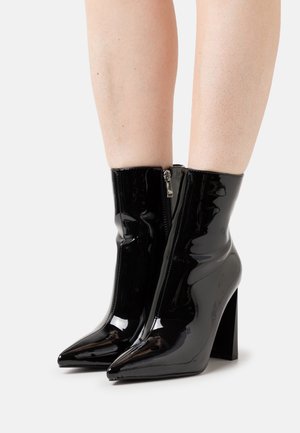 Botines de charol negro brillante con puntera afilada, tacones de bloque y cremalleras laterales, llevados sobre piernas desnudas.