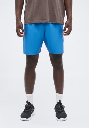 Hombre con pantalones cortos deportivos de color azul brillante, calcetines blancos y zapatos deportivos negros, de pie frente a un fondo liso y claro.