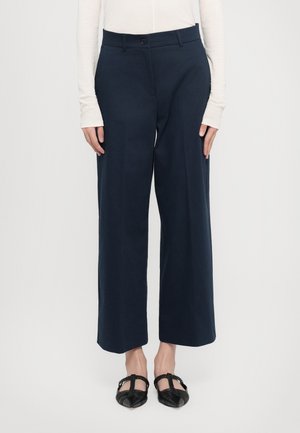 Femme portant un pantalon large bleu marine, un haut blanc à manches longues et des ballerines noires à bout pointu avec des brides tressées, debout devant un fond blanc.