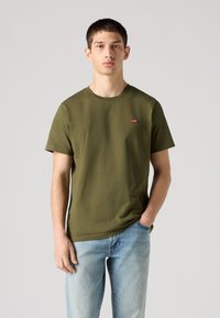 Oljčno zelena bombažna majica z okroglim izrezom, kratkimi rokavi in majhnim rdečim logotipom. Kombinirana z svetlo modrimi denim kavbojkami, stoječa pred nevtralnim ozadjem.