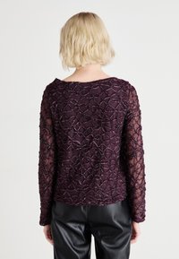 Bluza s dugim rukavima, ljubičasta, ukrašena perlama, s složenim cvjetnim čipkastim uzorkom, prozirnog materijala i okruglim izrezom. Uparena s crnim kožnim hlačama.