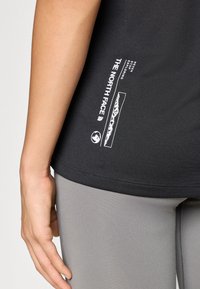 Μαύρο αθλητικό t-shirt με λευκό λογότυπο και γραφικό βουνού στο κάτω αριστερό μέρος, συνδυασμένο με λείες γκρι κολάν.