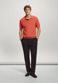 Jeune homme debout les mains dans les poches, portant un polo rouge à manches courtes, un pantalon noir et des chaussures habillées noires sur un fond uni.