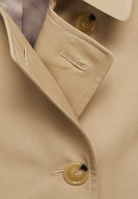 Détail d'un trench-coat beige avec un col pointu, une fermeture à gros boutons, et un tissu lisse avec une légère brillance.