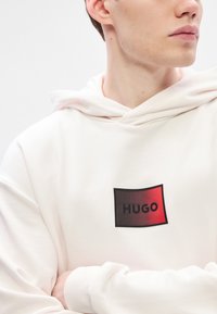 Λευκή κουκούλα με υφή, με κόκκινο και μαύρο λογότυπο που γράφει "HUGO." Διαθέτει κουκούλα με κορδόνι και ριμπ μανίκια. Μαλακή υφή υφάσματος.