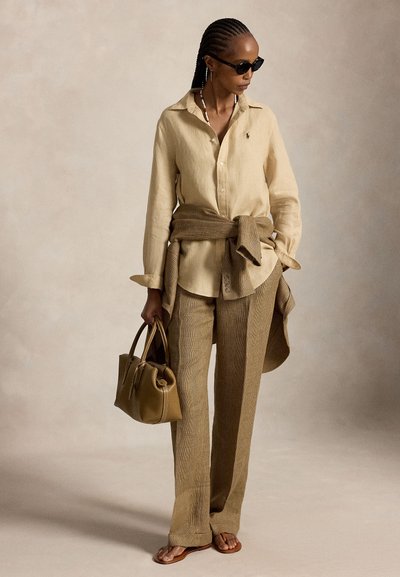 Femme portant une chemise beige à boutons, un pantalon à carreaux et des sandales, tenant un sac à main olive, avec une veste à carreaux nouée autour de la taille et des lunettes de soleil noires.