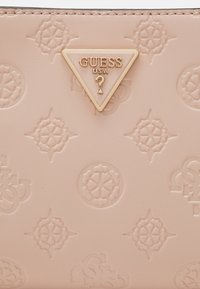 Cuir texturé rose clair avec logos embossés et un emblème triangulaire à bordure dorée portant l'inscription "GUESS USA ?" au centre.
