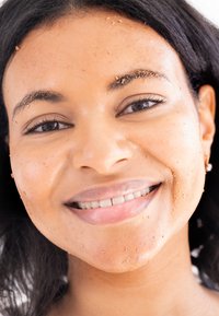 Nahaufnahme eines Gesichts mit einer hellbraunen Peelingmaske, das feuchte Haut mit kleinen Tropfen und natürlich dunklem Haar zeigt, wobei die Gesichtszüge hervorgehoben werden.