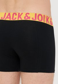 Černé boxerky s žlutým páskem, na kterém je růžový text "JACK & JOK." Tkanina vypadá hladce, s těsným střihem a čistými švy.