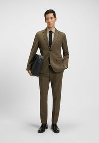 Completo marrone a righe con giacca aderente e pantaloni a sigaretta. Abbinato a una camicia verde chiaro, cravatta nera e una valigetta in pelle nera.