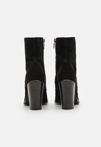 Paire de bottines en daim noir avec talons blocs et fermetures éclair à l'arrière, vues de dos sur fond blanc.