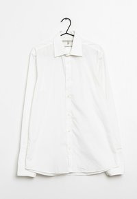 Camisa blanca de manga larga y botones colgada en una percha. Presenta un cuello clásico, botones en la parte delantera y un dobladillo ligeramente redondeado. Textura lisa y sencilla.