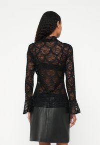 Lokkis juustega naine kannab pika varrukaga musta pitspluusi ja musta nahkseelikut, seistes seljaga kaamera poole.