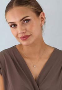 Goldene Halskette mit einem runden, klaren Anhänger. Das Model trägt eine taupefarbene Wickelbluse. Schlichte Steckerohrringe ergänzen den Schmuck. Heller Hintergrund.