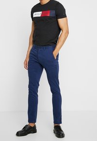 Man som bär en svart kortärmad T-shirt med "Hilfiger"-logotyp, blå slim fit-byxor och svarta snörskor, står med en hand i fickan.