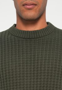 Maglione a maglia verde oliva con collo rotondo a coste e motivo testurizzato, caratterizzato da un design a griglia su tutta la superficie.