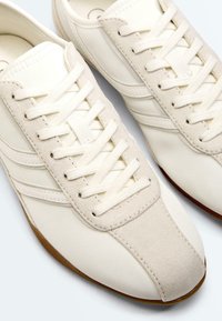 Gros plan sur des baskets blanc cassé avec des embouts en daim, des parties supérieures en tissu, des lacets assortis et des semelles en gomme.