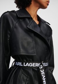 Melns mākslīgās ādas tranče mētelis ar platām apmalēm, savilkta vidukļa un josti, uz kuras ir balts teksts "KARL LAGERFELD JEANS".