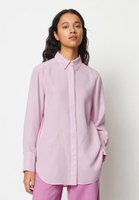 Camicia a maniche lunghe rosa chiaro con colletto abbottonato, taglio dritto e spacchi laterali, realizzata in un tessuto morbido. Abbinata a pantaloni rosa.