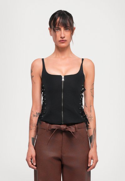 Femme aux cheveux bruns portant un haut sans manches noir avec fermeture éclair à l'avant et un pantalon taille haute marron avec une ceinture nouée, se tenant devant un fond blanc.