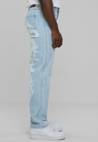 Hombre que lleva unos pantalones vaqueros de un azul claro con impresiones tribales blancas en el lateral y zapatillas Nike Air Force 1 blancas, sobre un fondo gris.