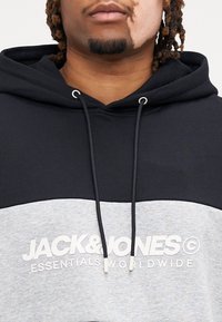 Capuchon trui met zwart bovenstuk en grijs onderpaneel. Heeft een witte bedrukte logo "JACK & JONES ESSENTIALS WORLDWIDE" en trekkoordaccenten.