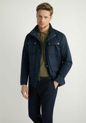 Młody mężczyzna z jasnobrązowymi włosami, ubrany w granatową kurtkę na oliwkowej koszulce polo i czarne spodnie, stoi na tle prostego jasnego tła.