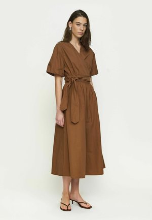 Young woman with long brown hair wearing a brown midi wrap dress with wide sleeves and tied waist, standing with neutral expression and heeled sandals.
