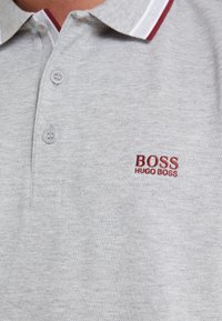 Polo gris clair avec deux boutons et un col orné d’une fine rayure rouge et blanche, brodé du logo rouge « BOSS HUGO BOSS ».
